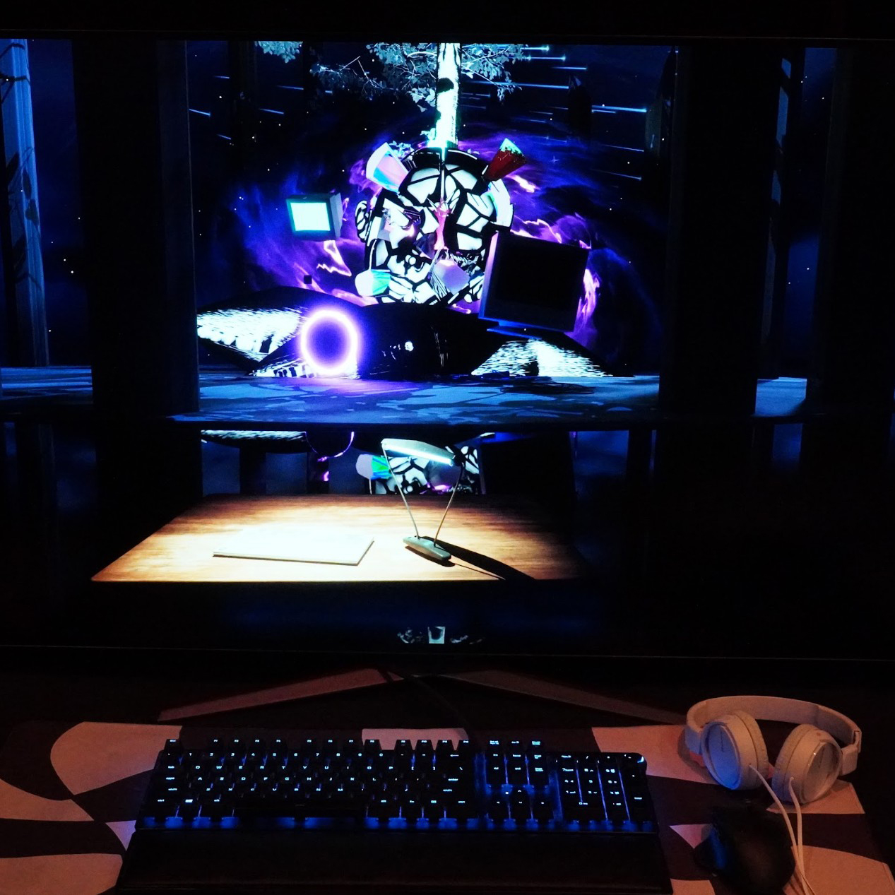 A dark installation view shows a computer monitor displaying a vivid Unreal Engine environment with glowing purple and blue lighting, abstract structures, and floating digital forms. In front of the screen are a backlit keyboard, mouse, and headphones, suggesting an interactive virtual dream world experienced through sound, navigation, and immersive visuals.