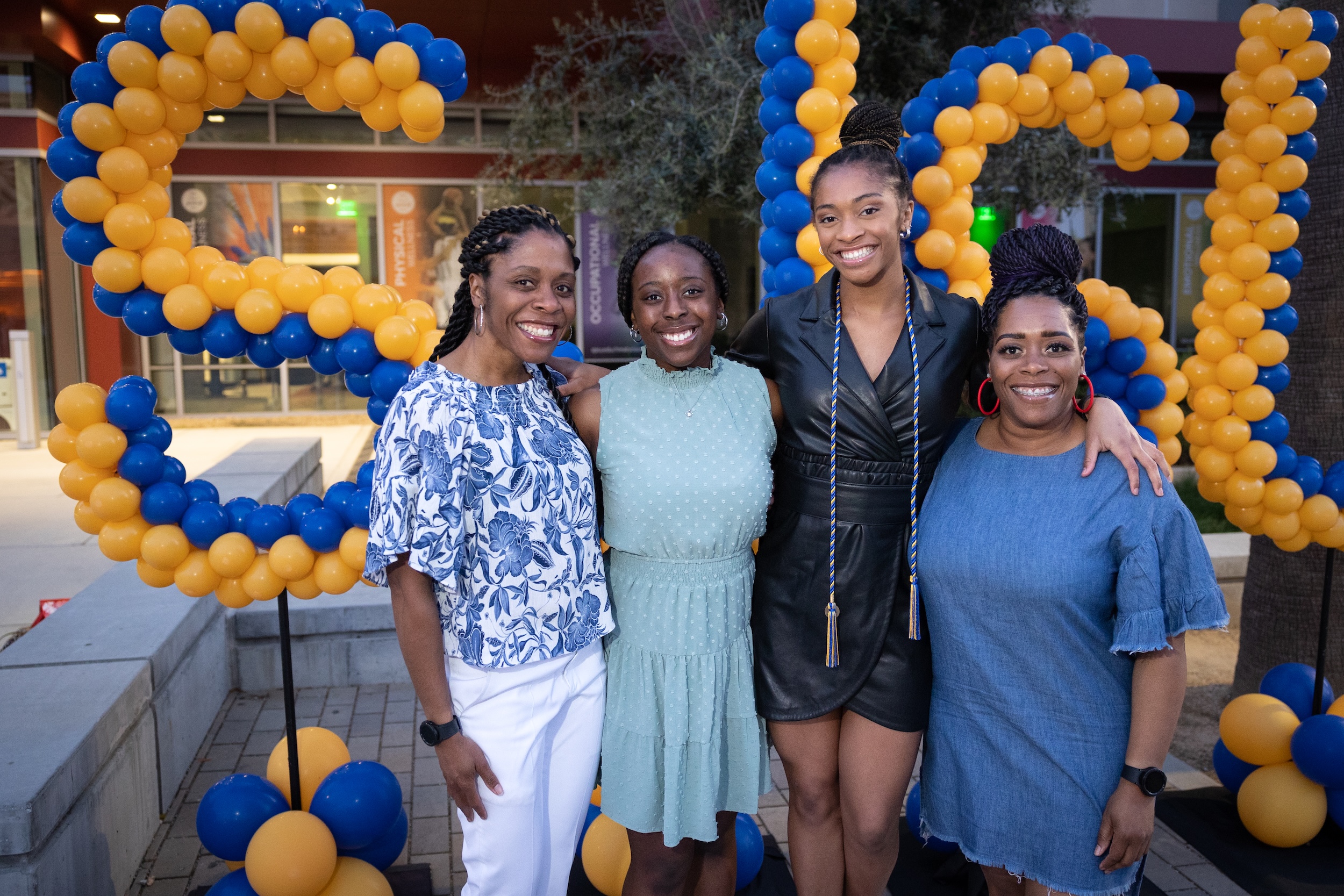 Four individuals smiling and posing together in front of large blue and gold balloon decorations. The festive setting suggests a celebratory event, with two of the individuals wearing honor cords.