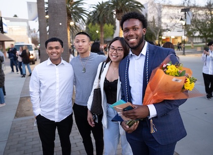 Four young adults smiling and posing for a photo outdoors in the evening. One person on the right is dressed in a suit with honor cords and holding a bouquet of flowers. The other three are casually dressed. Palm trees and campus buildings are visible in the background, with other people walking and gathering nearby."