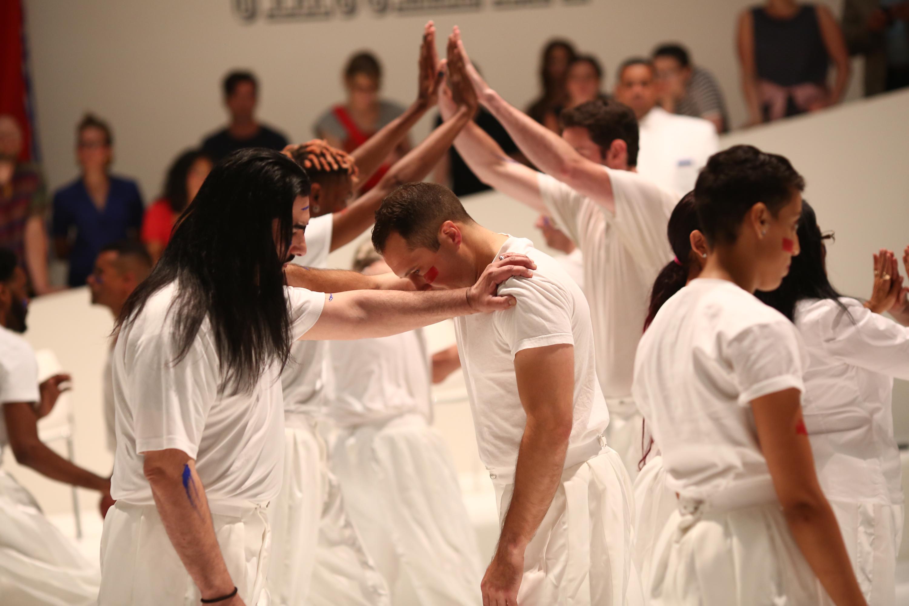 Image caption: Primitive Games - performance, 1 hr. at Guggenheim Museum, New York, NY, 6/21/18. Photo by Paula Court performance still of figures wearing white clothing and joining hands. The two figures in the foreground rest their hands on each other shoulders